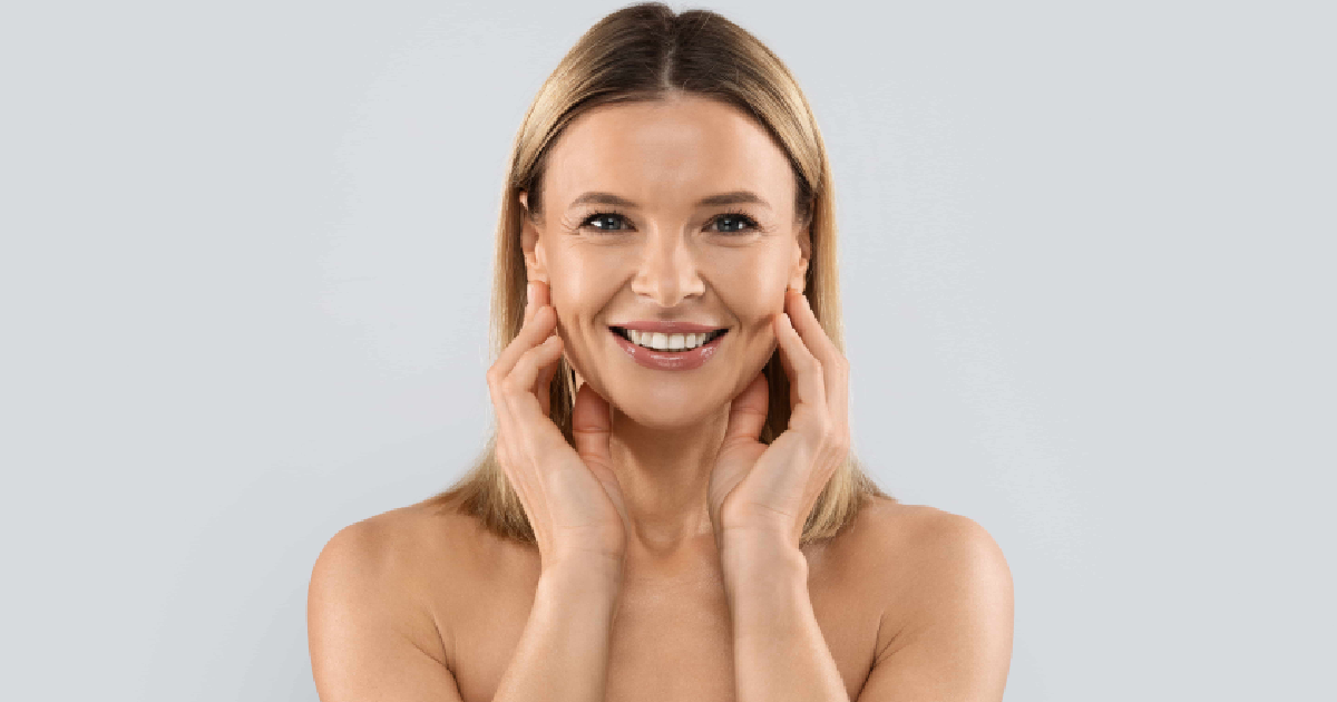 Blonde woman with a bob smiles, hands on cheeks, wearing white towels, representing Sculptra in Edina, MN, against a light gray background.