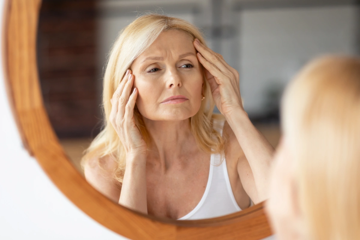 Woman examining fine lines while looking in a mirror, illustrating how HydraFacial supports smoother, hydrated, and firmer-looking skin.